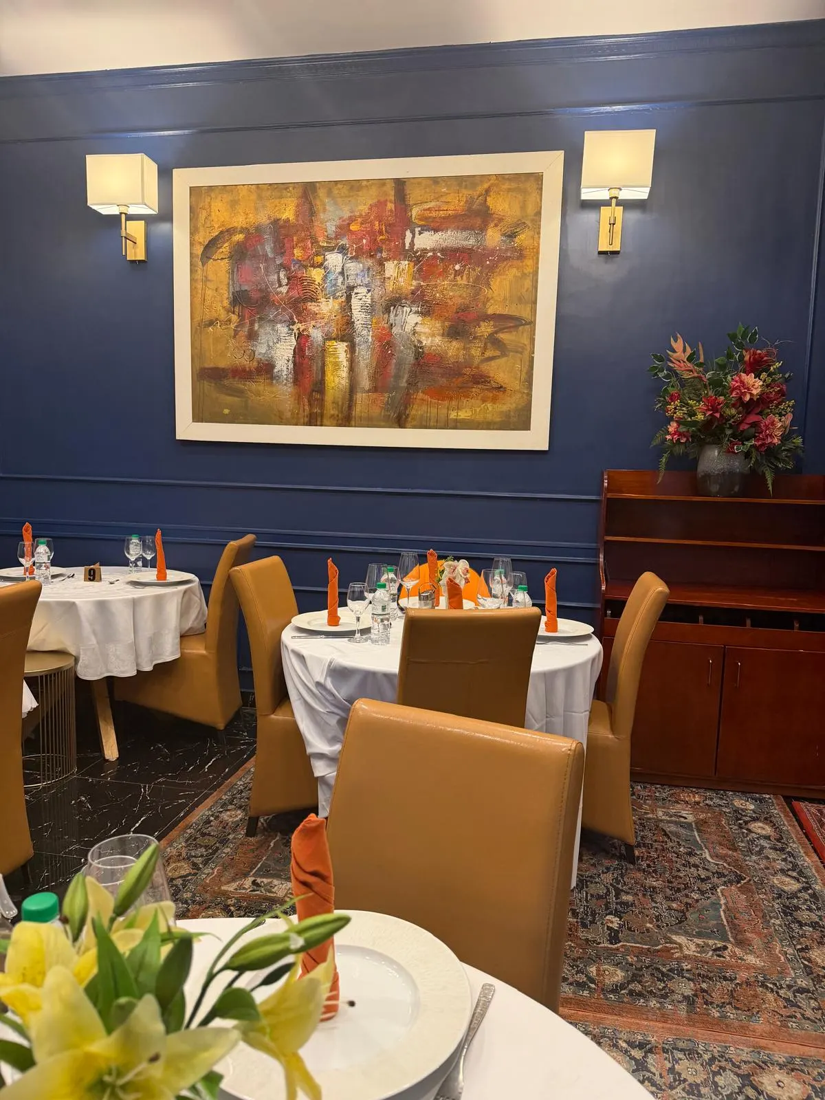 Salle aux murs bleus avec grande toile abstraite, tables rondes nappées et buffet en bois surmonté d un bouquet