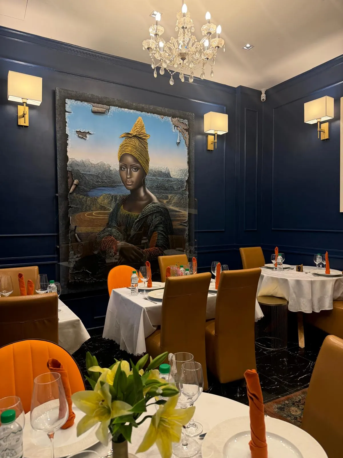 Salle du restaurant aux murs bleu nuit, grand portrait peint d'une femme à la coiffe dorée, tables dressées de nappes blanches et serviettes orange